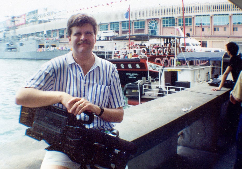 A man sitting on a waterfront ledge in Hong Kong with a broadcast camera resting in his lap, with boats and a busy harbor building visible in the background.